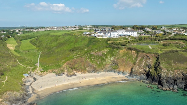 Vue aérienne d’une zone côtière avec une plage de sable, des eaux turquoise et limpides, et un grand bâtiment blanc en arrière-plan.