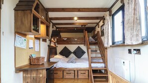 A compact, rustic cabin-style room with a bunk bed, wooden beams, a small kitchenette, and a window with floral curtains. - Daisy (Woodbridge)