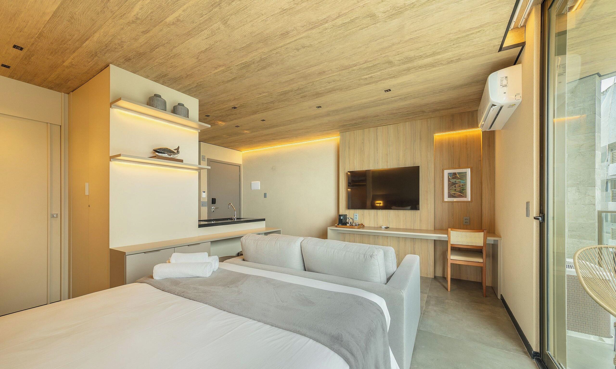 A modern hotel room with a wooden ceiling, a bed with white bedding, a desk with a flat-screen TV, and a seating area with a chair.