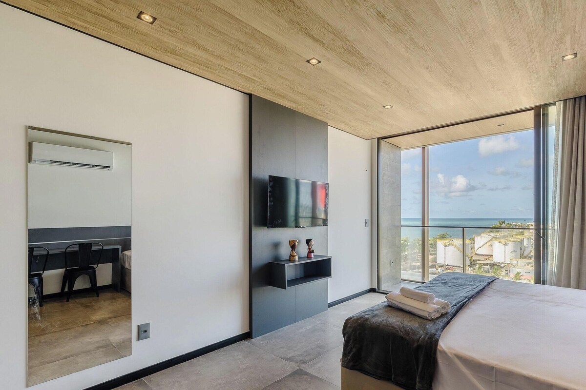 Een moderne hotelkamer met een groot bed, een flatscreen-tv aan de muur, een klein bureau met een stoel, en een balkon met uitzicht op de stad en de zee.
