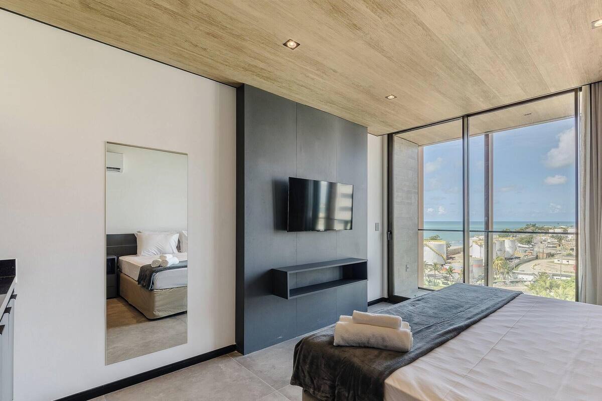 A modern bedroom with a large bed, a flat-screen TV mounted on the wall, and a balcony with a city view.