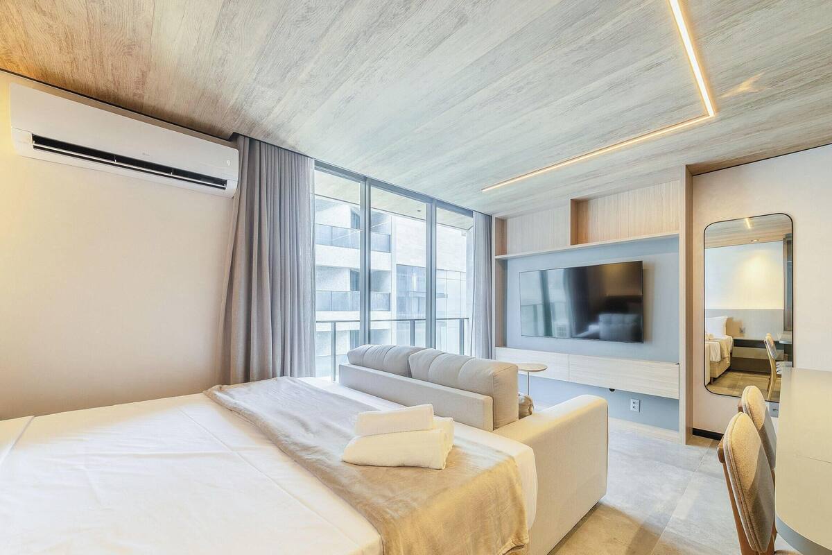 A modern hotel room with a large bed, a sofa, a flat-screen TV mounted on the wall, and a large window offering a view of the city.