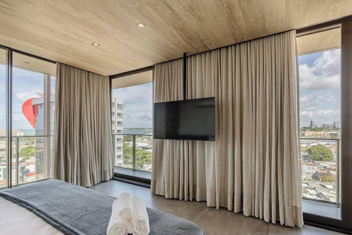 A modern bedroom with a large bed, a flat-screen TV, and floor-to-ceiling windows offering a city view.
