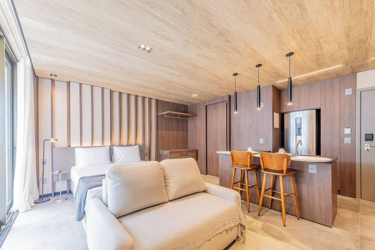 A modern hotel room with a sofa, a dining area with stools, a kitchen with a refrigerator, and a large window.