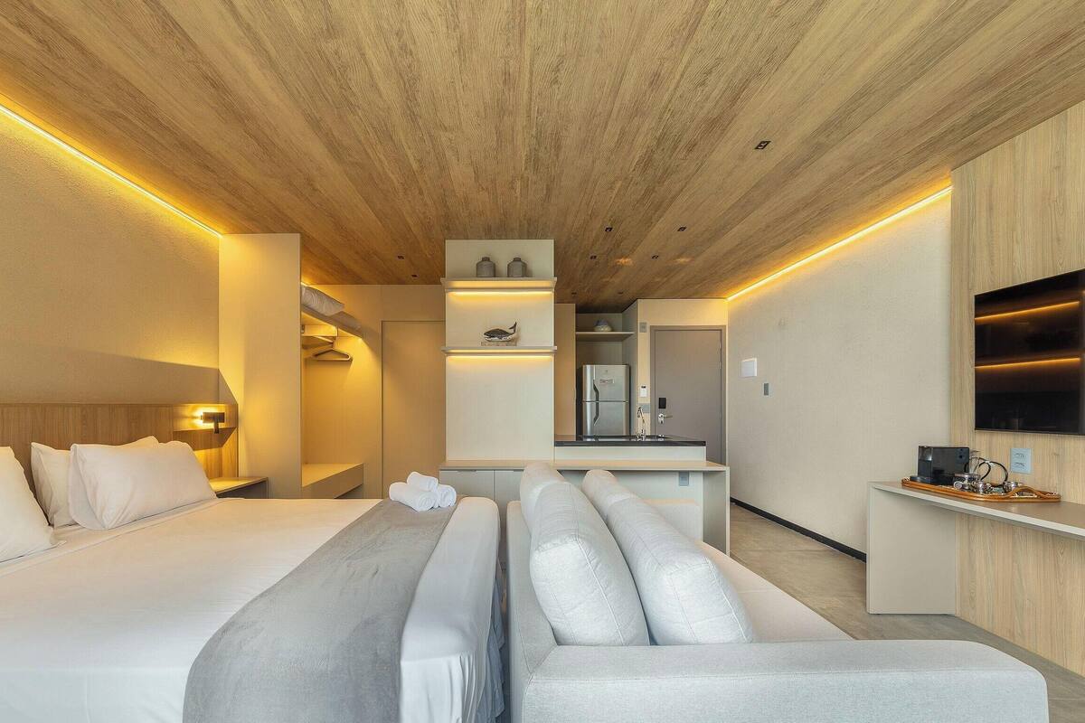 A modern hotel room with a wooden ceiling, a bed with white linens, a seating area with a grey cushion, a small kitchenette, and a flat-screen TV mounted on the wall.
