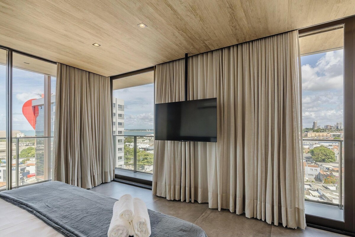 Een moderne slaapkamer met een groot bed, een flatscreen televisie en raam-tot-plafond ramen met uitzicht op de stad.