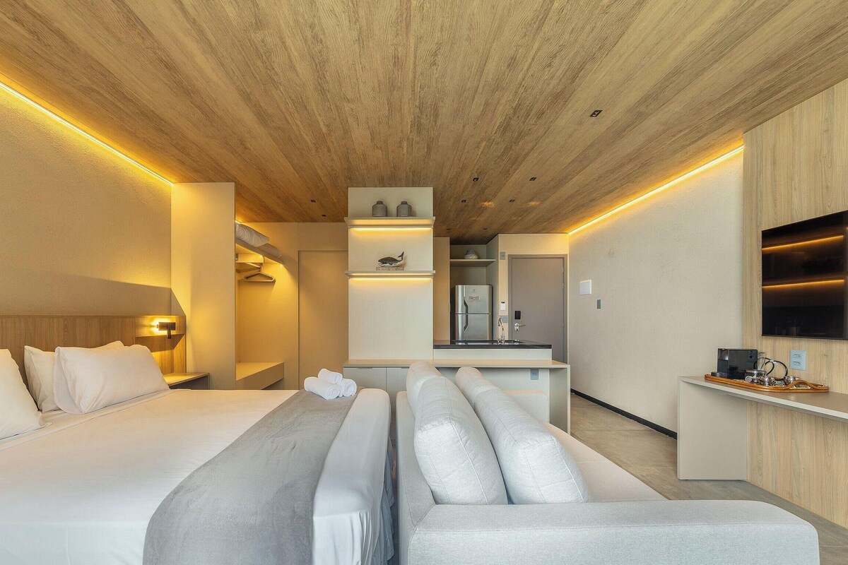 Een moderne hotelkamer met een houten plafond, een bed met witte lakens, een zithoek met een grijze kussen, een kleine kitchenette en een flatscreen televisie aan de muur.