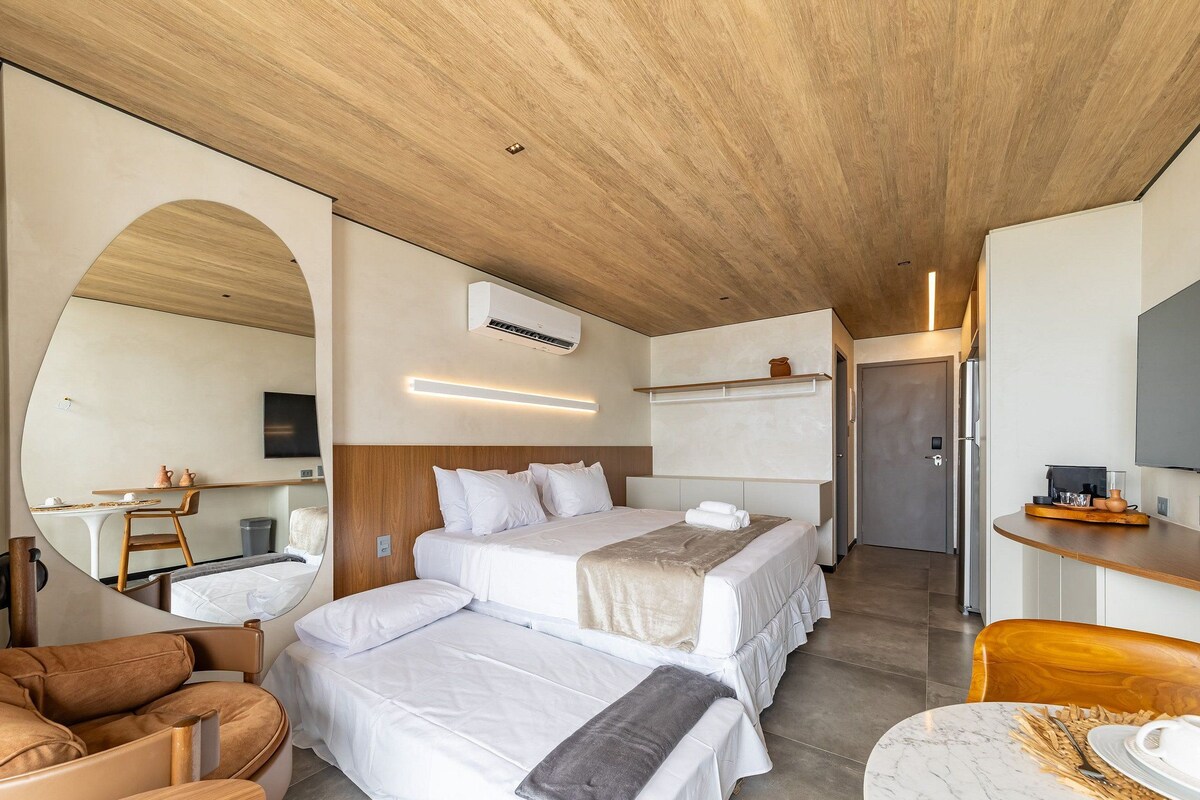 A modern hotel room with a wooden ceiling, a large mirror, a bed with white bedding, a desk with a flat-screen TV, and a seating area with a leather chair.