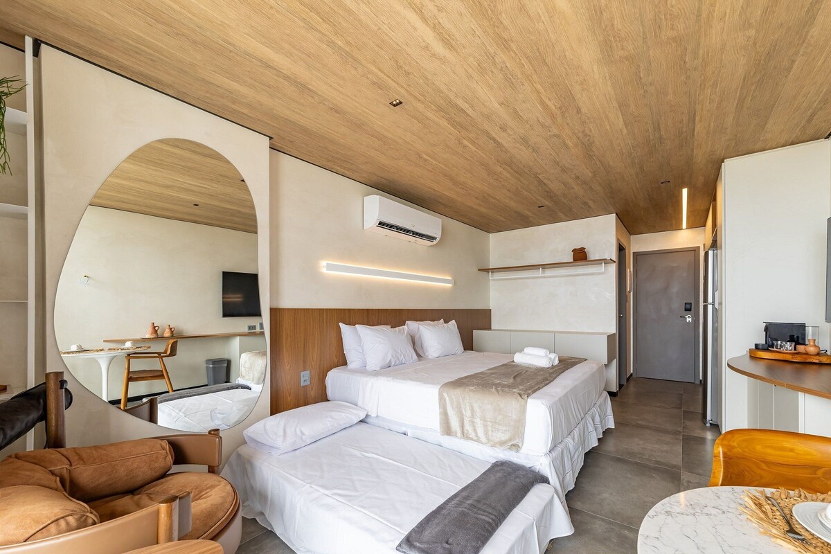 Een moderne hotelkamer met een houten plafond, een grote spiegel, een bed met witte beddengoed, een bureau met een flatscreen televisie en een zithoek met een lederen fauteuil.