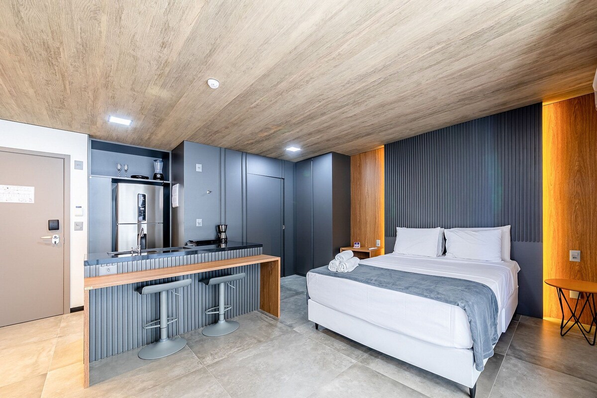 Een moderne hotelkamer met een houten plafond, een bed met wit beddengoed, een geïntegreerde keuken met een koelkast, een koffieapparaat en twee barkrukken.