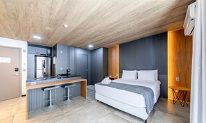 Een moderne hotelkamer met een houten plafond, een bed met wit beddengoed, een geïntegreerde keuken met een koelkast, een koffieapparaat en twee barkrukken.