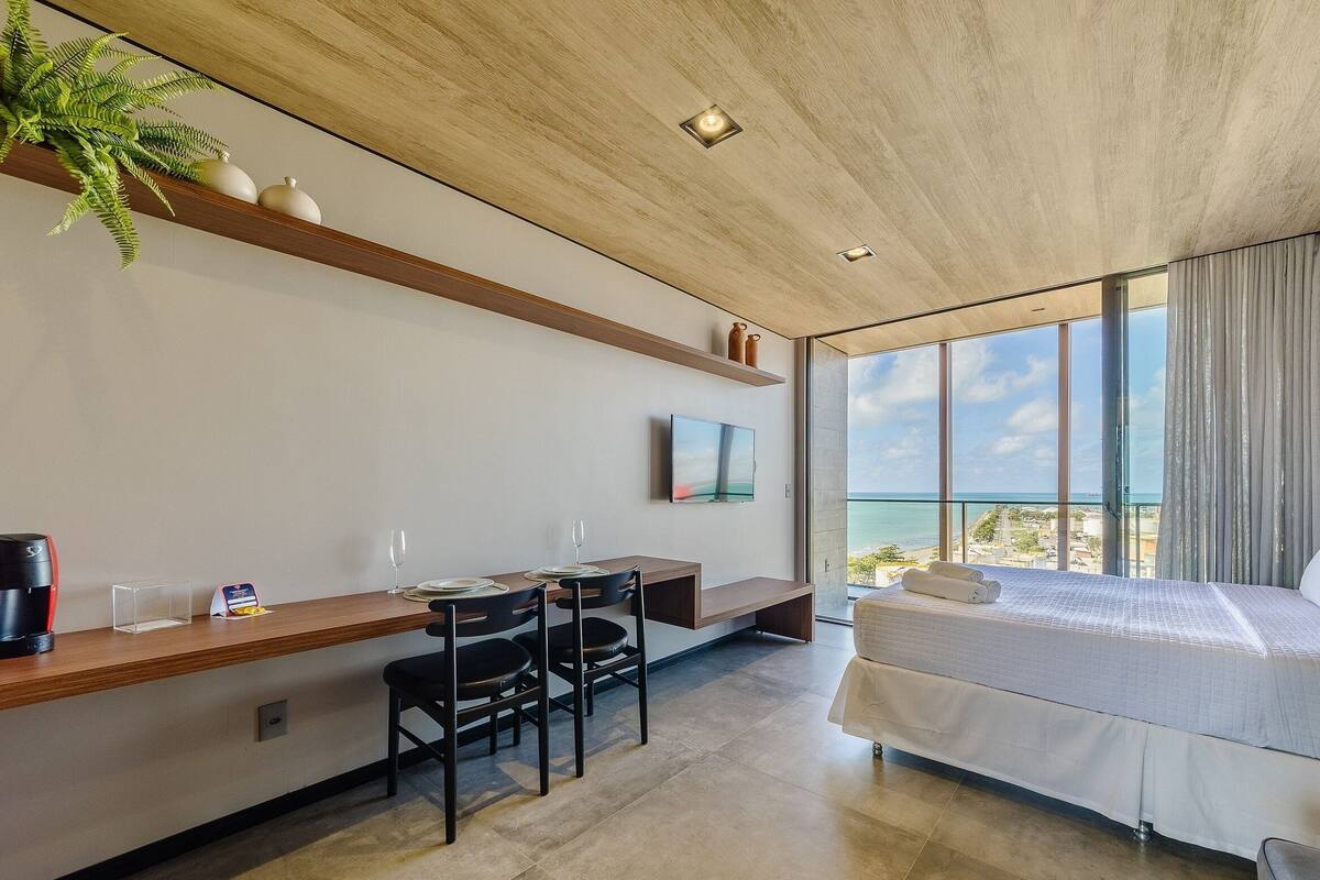 A modern hotel room with a large bed, a desk with chairs, a TV, and a view of the sea.
