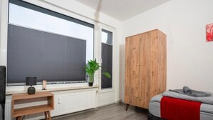 A bedroom with a bed, a wooden wardrobe, a radiator, a window with blinds, a small table with a lamp, and a plant.