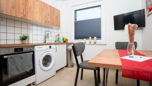 A modern kitchen with wooden cabinets, a washing machine, a dining table with chairs, and a TV mounted on the wall.