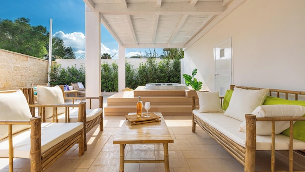Un espacio al aire libre cubierto con mobiliario de madera y una mesa central, con vistas a un jardín y una bañera de hidromasaje.