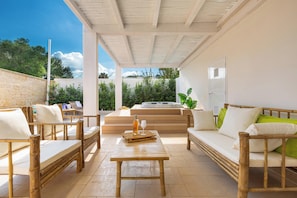 Un espace extérieur aménagé, abrité et doté de mobilier en bois, avec une table centrale, offrant une vue sur un jardin et un jacuzzi.