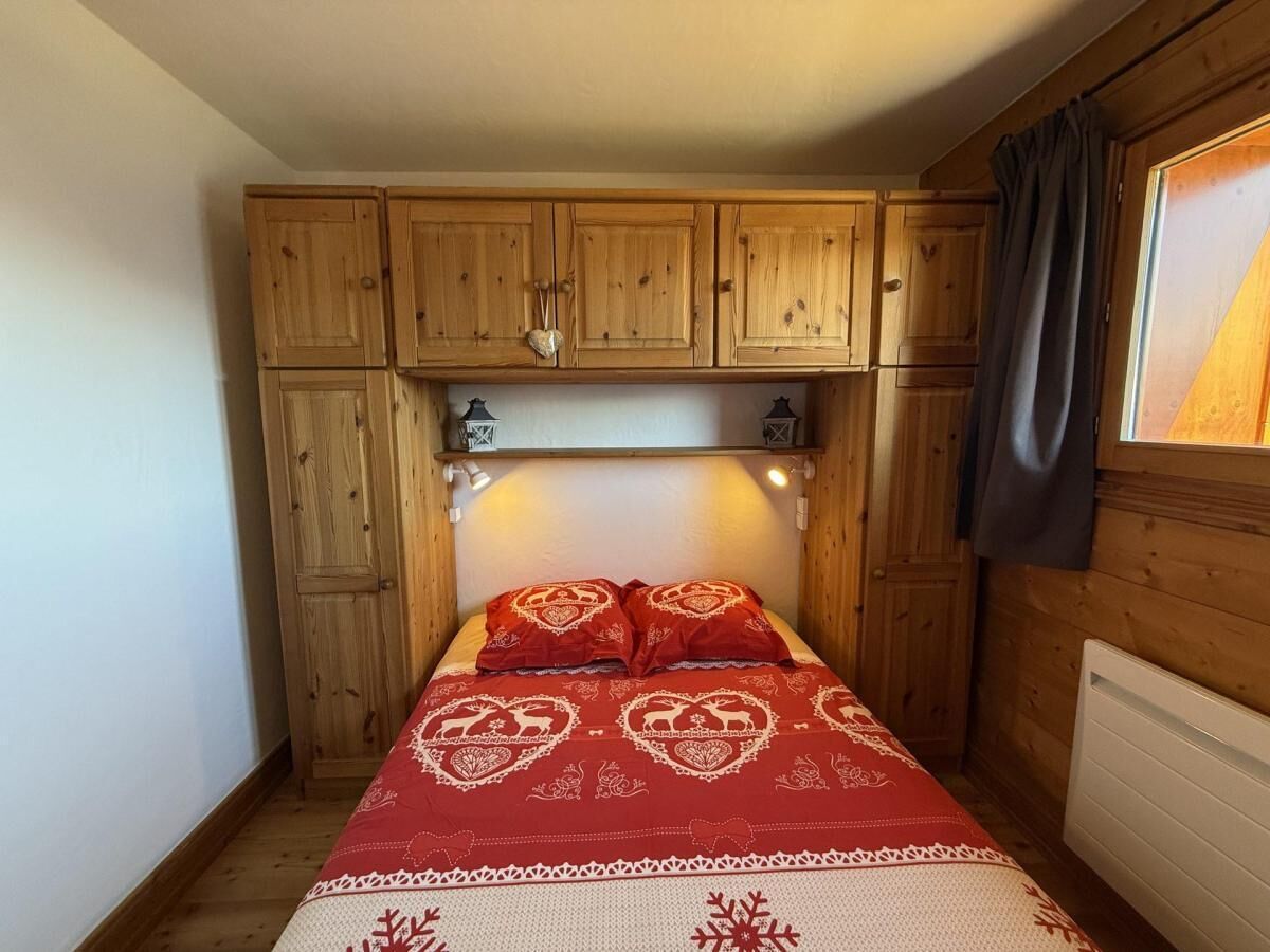 A small bedroom with wooden cabinets above the bed, a red bedspread with heart patterns, and a window with curtains.