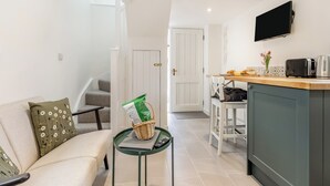 A compact living space with a sofa, a small table with a basket, a kitchenette with a TV, and a door leading to another room. - The Little House - UK49129 (Stapleton)
