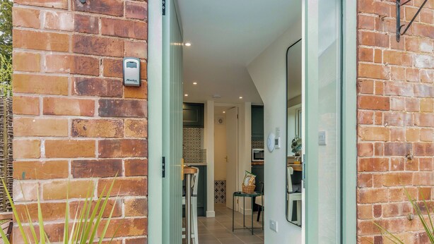 A narrow hallway with a brick wall, a door leading to a room with a microwave, and a small table with a basket. - The Little House - UK49129 (Stapleton)