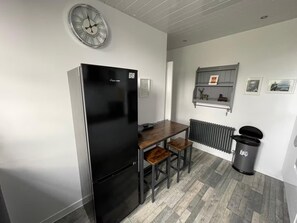 A compact kitchen with a black refrigerator, a wooden table with two chairs, a wall-mounted clock, and a radiator. - Isle of Bute Blissful Retreat- Stunning 3 Bed Flat (Rothesay)
