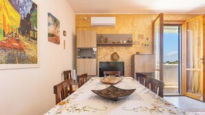 Un comedor con una mesa lista para cuatro personas, rodeada de sillas de madera. A la izquierda, un cuadro grande en la pared, y un estante con objetos decorativos sobre la mesa. Se ve el exterior a través de una puerta abierta.