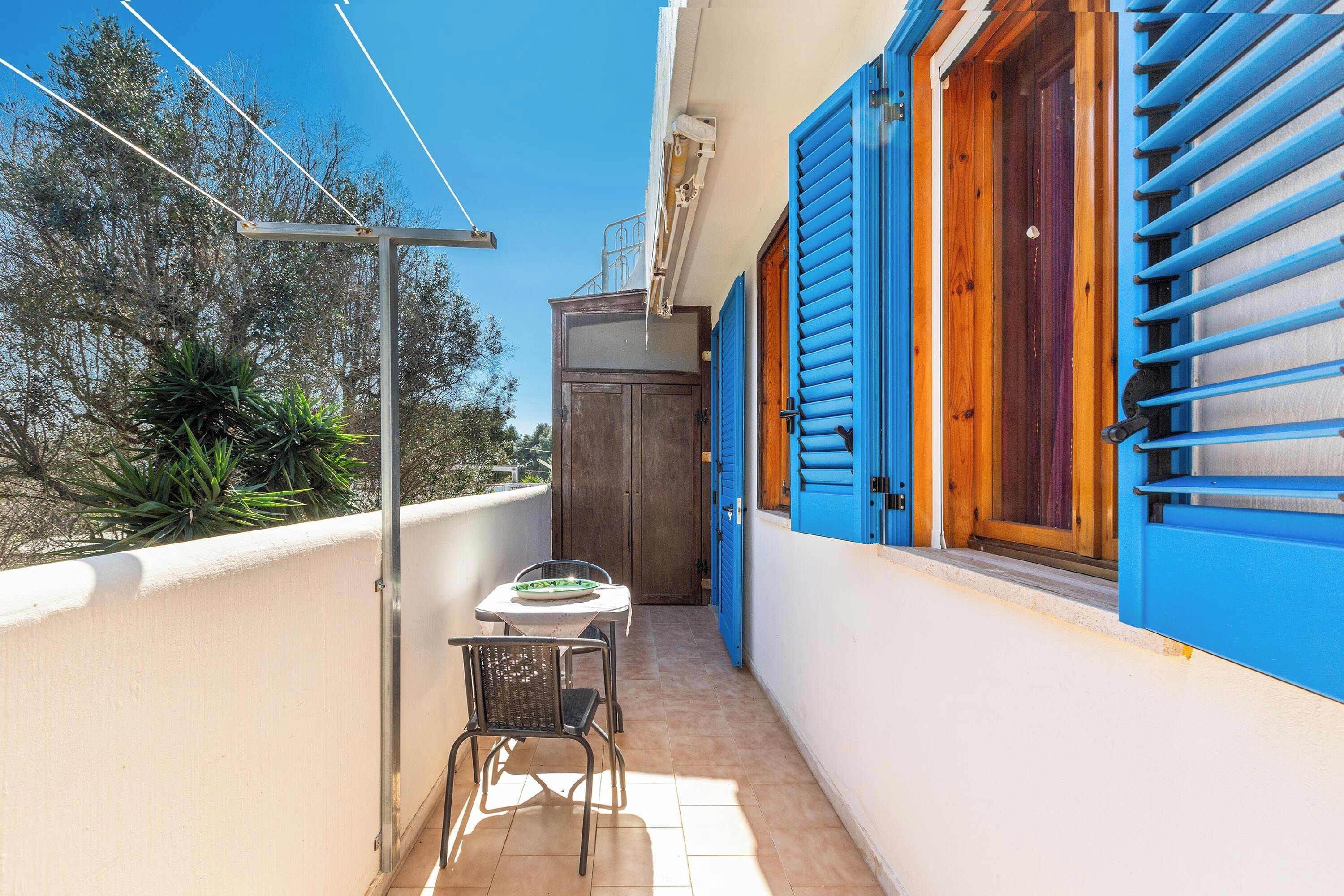 Een smal balkon met een tafel en stoelen, een houten deur en blauwe luiken.