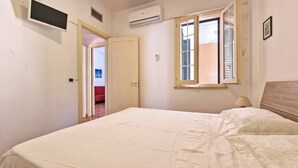Quarto com cama, janela com persianas, ar-condicionado e porta para outro cômodo.
