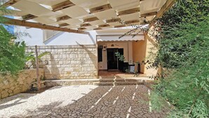 Área externa coberta com paredes de pedra e piso de azulejos, que dá acesso à entrada de um edif ício.