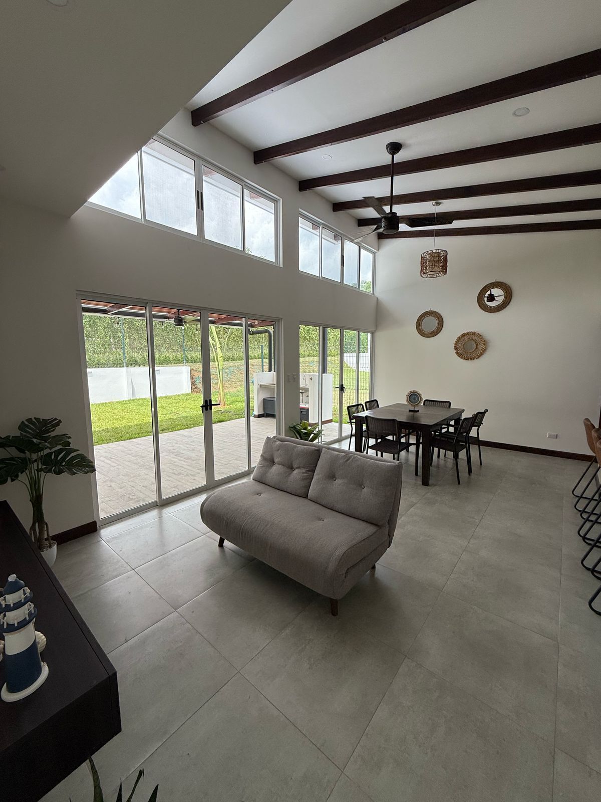 A spacious living area with a sofa, dining table, and chairs. Large windows provide natural light and views of the outdoors.