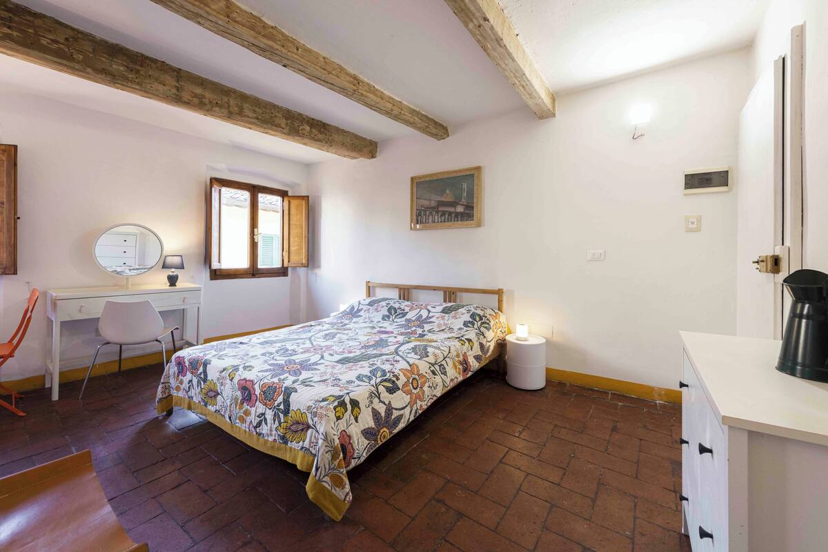A bedroom with a bed, a desk, a chair, a bedside table with a lamp, and a window.