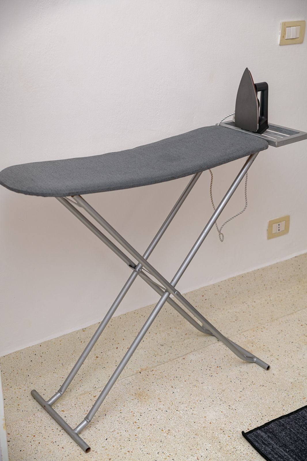 A portable ironing board with an iron, set against a plain wall with a switch and a power outlet.