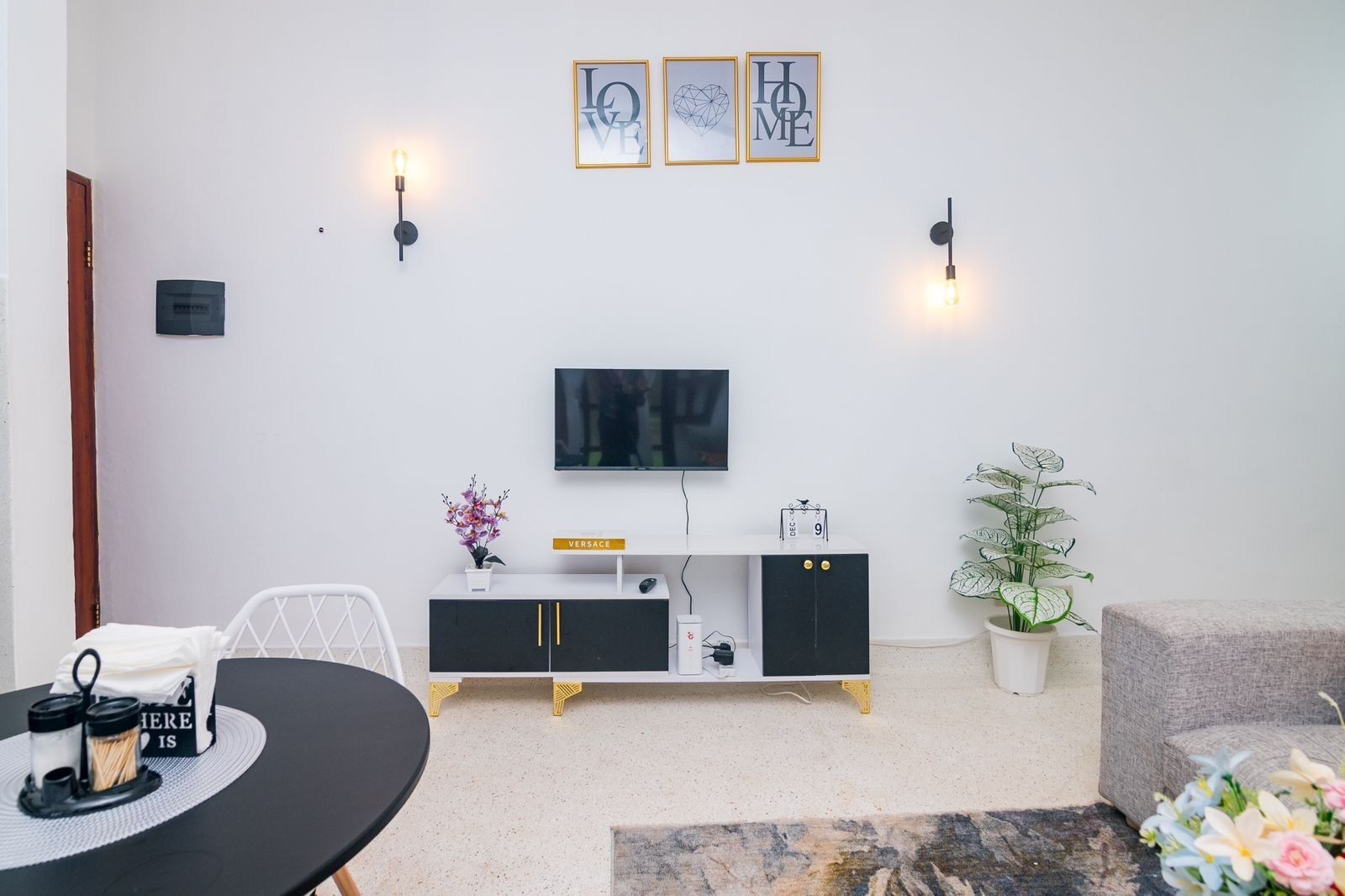 Un salon moderne avec un téléviseur à écran plat, un meuble noir et doré, une table de salle à manger ronde et une plante en pot.