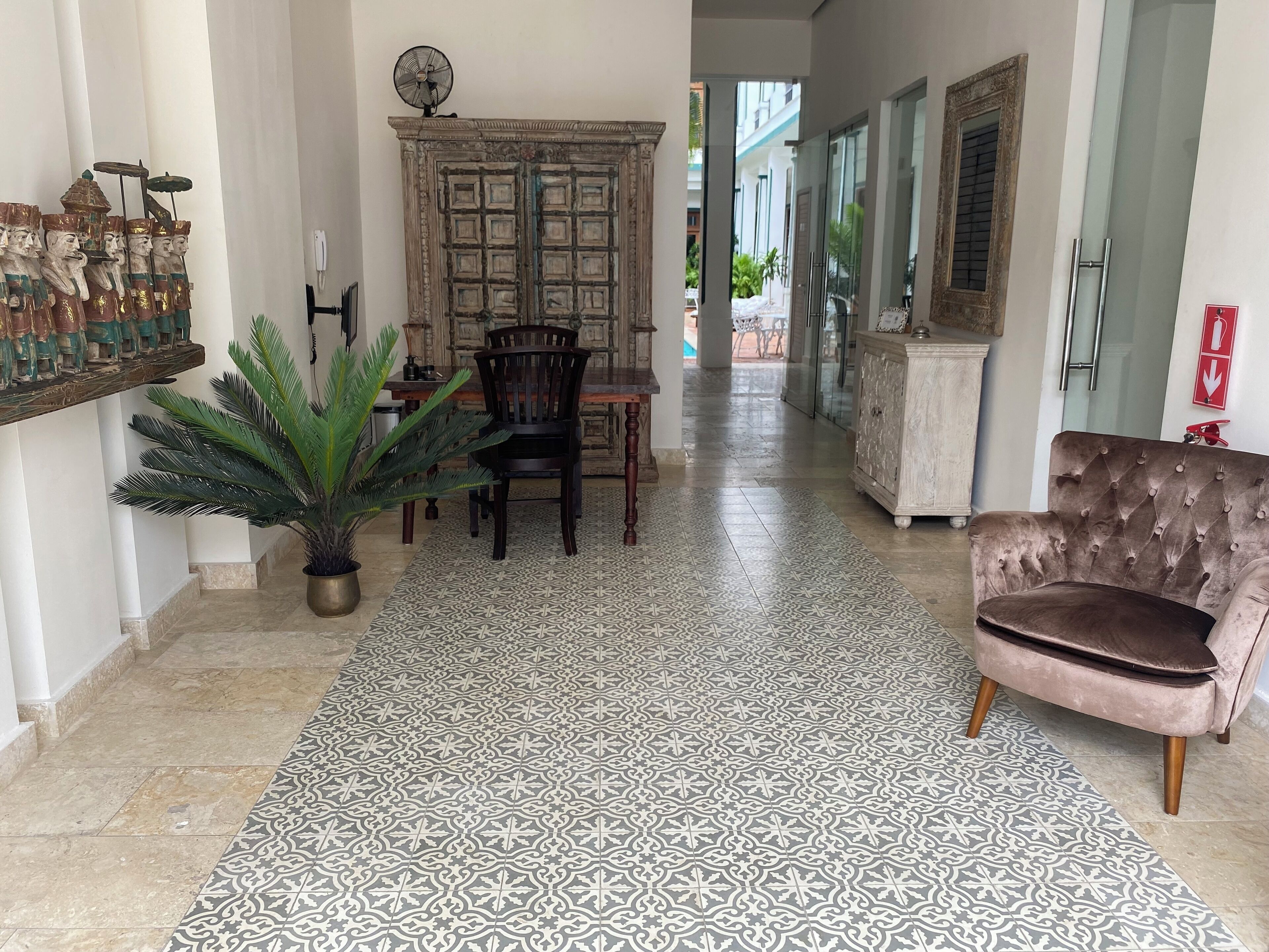 A hallway with patterned flooring, a dining area with chairs and a table, a potted plant, and a tufted armchair.