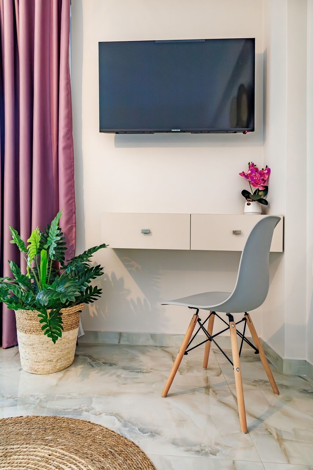 A modern living room with a flat-screen TV mounted on the wall, a grey chair with wooden legs, a small potted plant, and a woven rug on a marble floor.