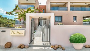 A modern building with a sign reading "Casa Helme," featuring a staircase leading to an upper floor, a small garden area with rocks and a potted plant, and a clear sky in the background. - Casa Helone  (Kassandra)
