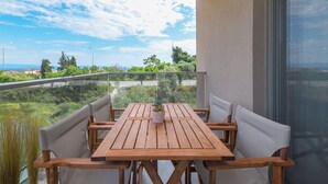 A wooden table with four chairs on a balcony with a glass railing, overlooking a green landscape and a clear sky. - Casa Helone  (Kassandra)