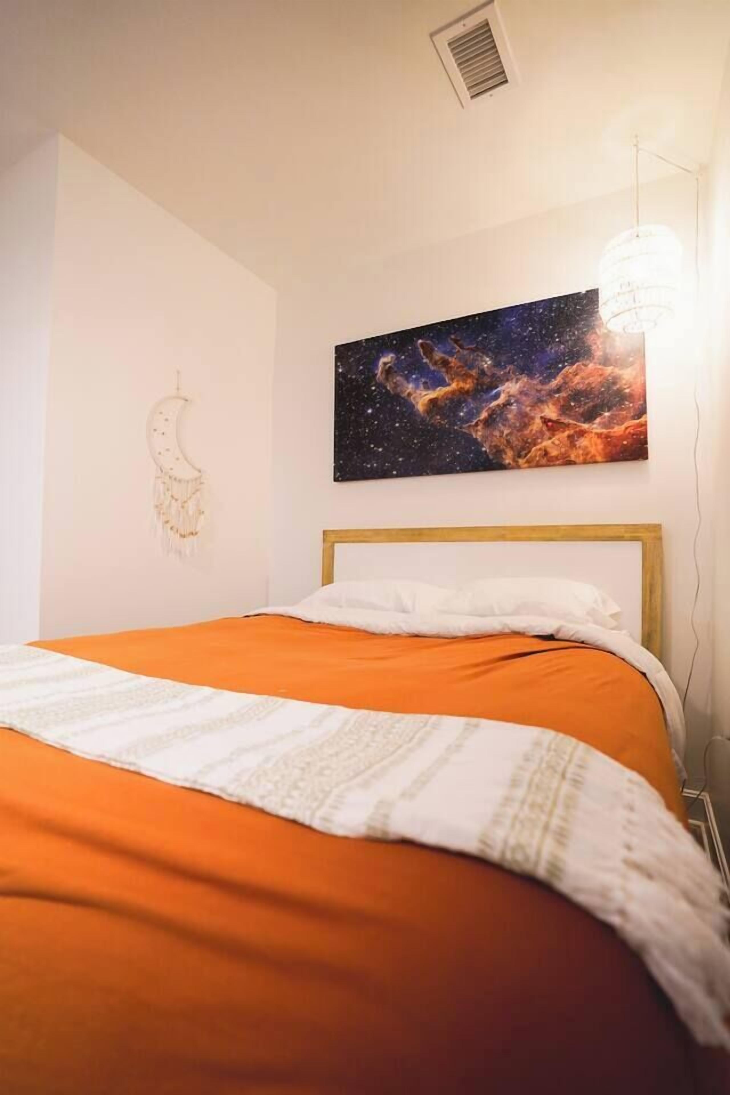 A bedroom with a bed, a wall-mounted painting of a nebula, a dreamcatcher, and a pendant light.