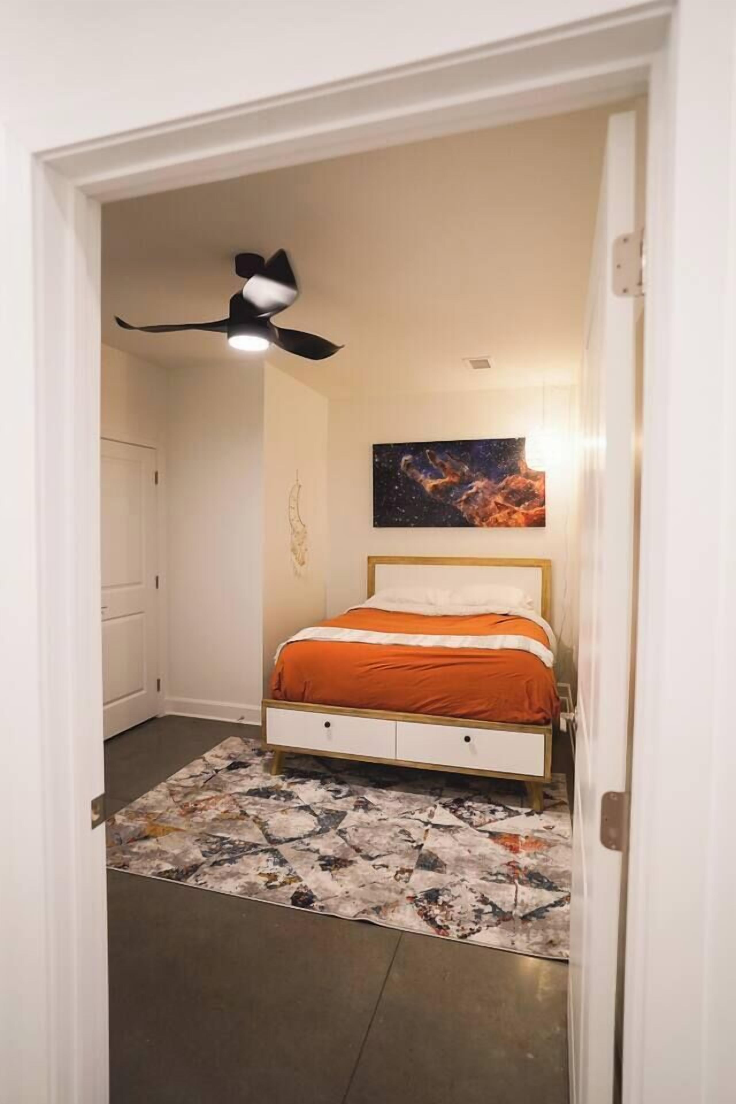 A bedroom with a bed, a ceiling fan, a painting, and a patterned rug.