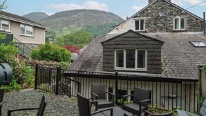Une maison avec une cheminée en pierre, une petite fenêtre et une terrasse en gravier agrémentée de chaises en osier.