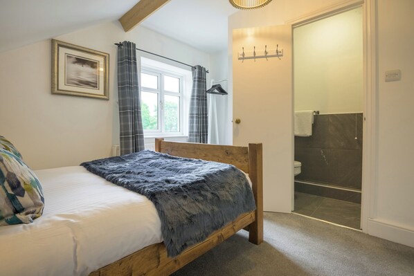 A bedroom with a wooden bed, a window with curtains, a framed picture on the wall, and a bathroom visible through an open door. - Moss Crag (Penrith)
