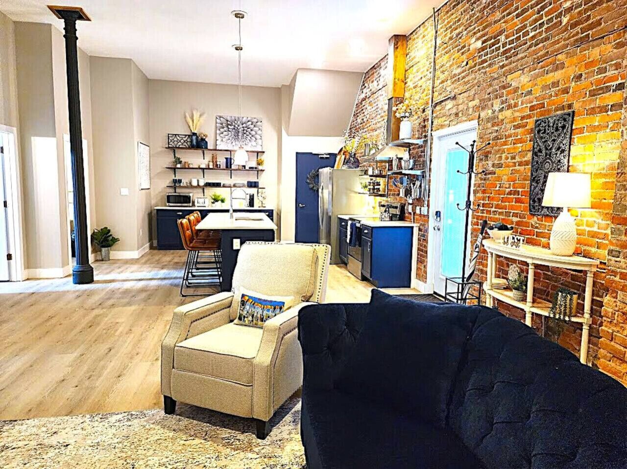 Una sala de estar con un sofá, una silla, un comedor con mesa y sillas, una cocina con refrigerador y armarios, y una pared de ladrillo.