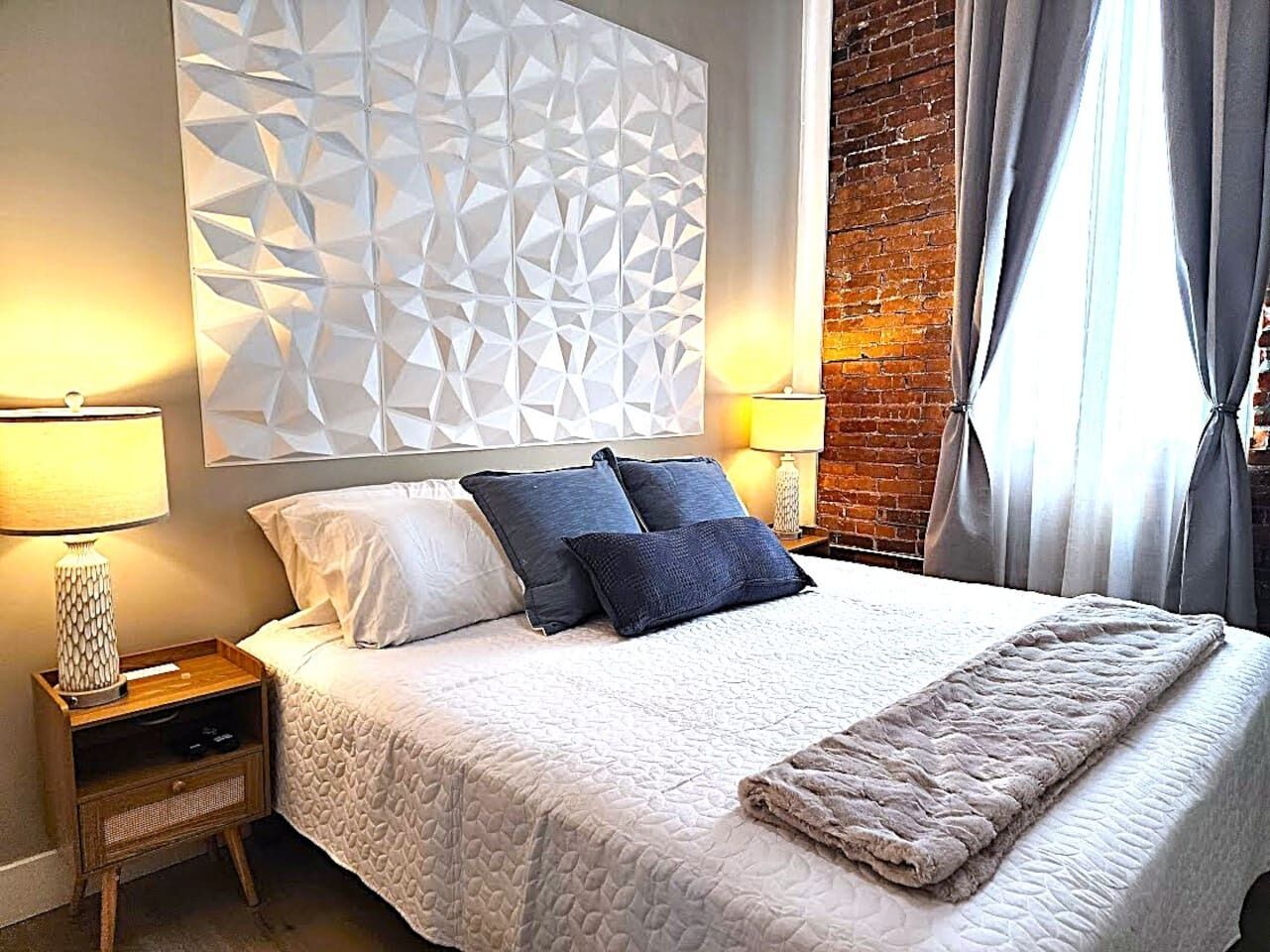 A neatly made bed with a white quilt, two pillows, a bedside table with a lamp, and a geometric wall panel.