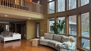 Amplia sala de estar con techo alto, ventanas grandes, escalera de madera empotrada y vistas al exterior.