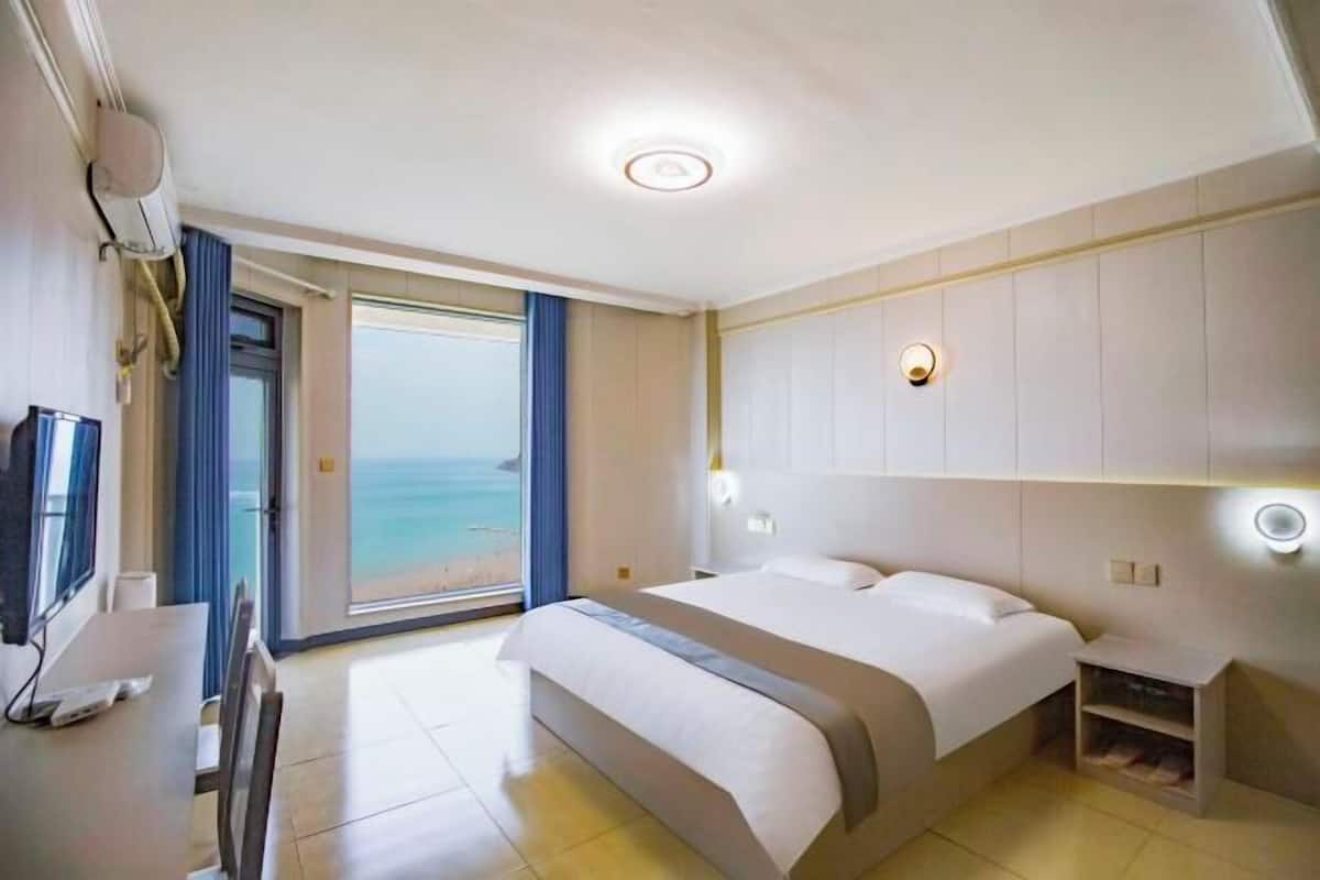 A hotel room with a bed, a desk, a chair, a TV, a bedside table, and a view of the ocean.
