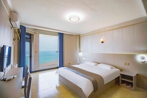 Habitación de hotel con cama, escritorio, silla, televisor, mesita de noche y vista al océano.