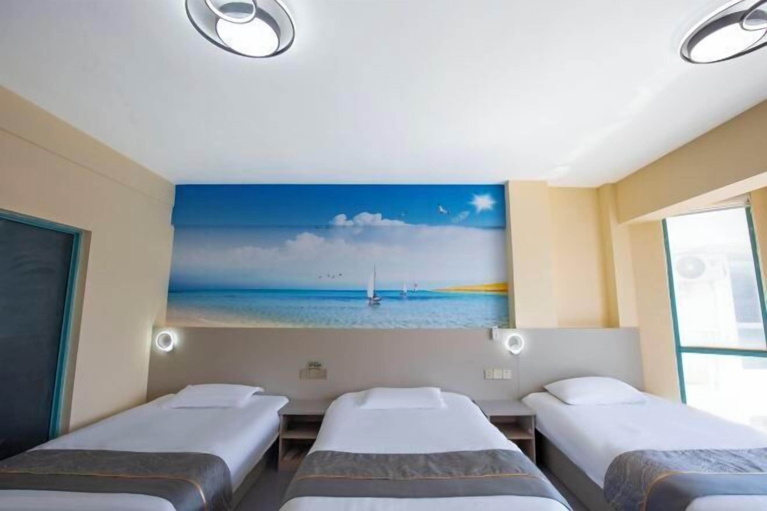A hotel room with three beds, a painting of a beach scene, and a window with a view of the sea.