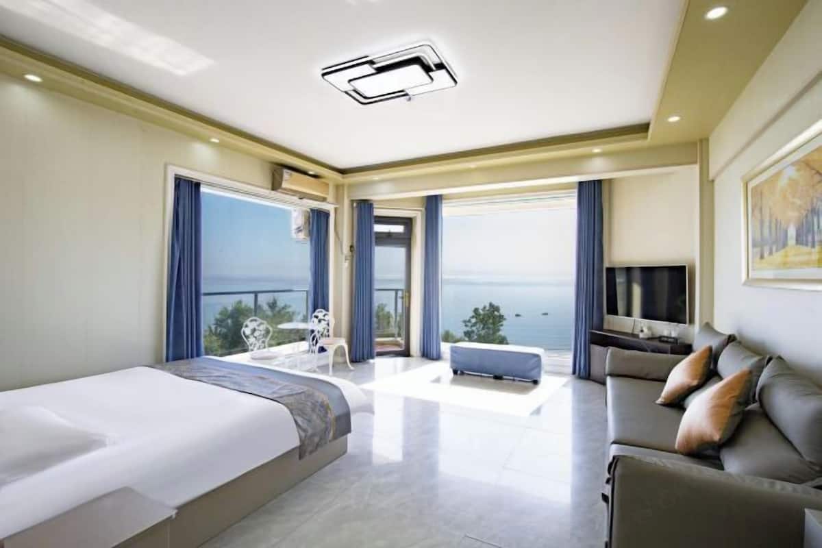 A modern hotel room with a large bed, a sofa, a TV, and a balcony with a view of the sea.