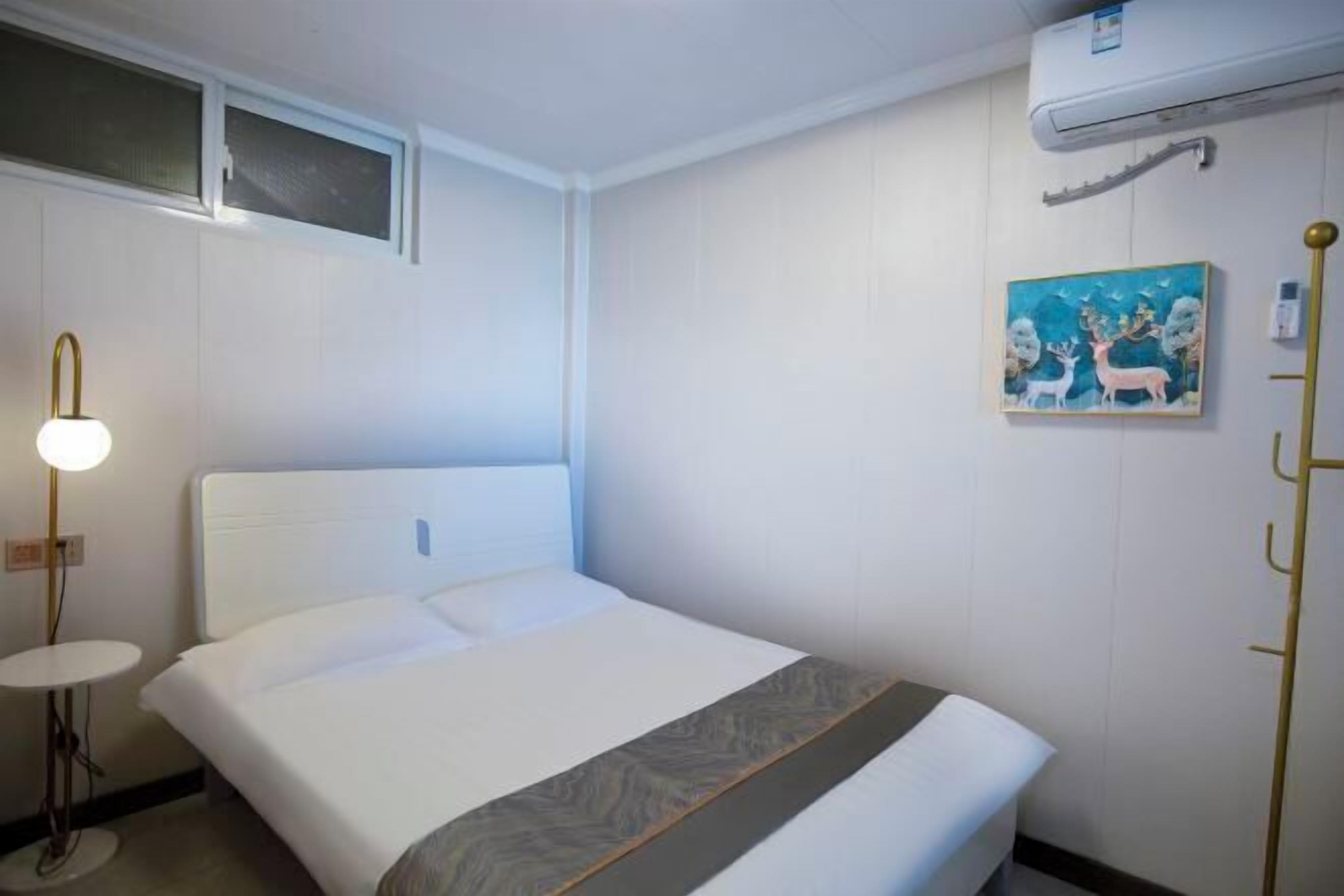 A hotel room with a bed, a bedside table, a wall-mounted air conditioner, and a painting.
