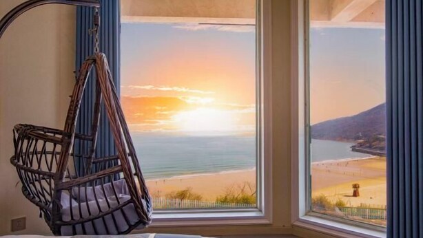 Quarto com uma grande janela que oferece vista para a praia e oceano ao pôr do sol.