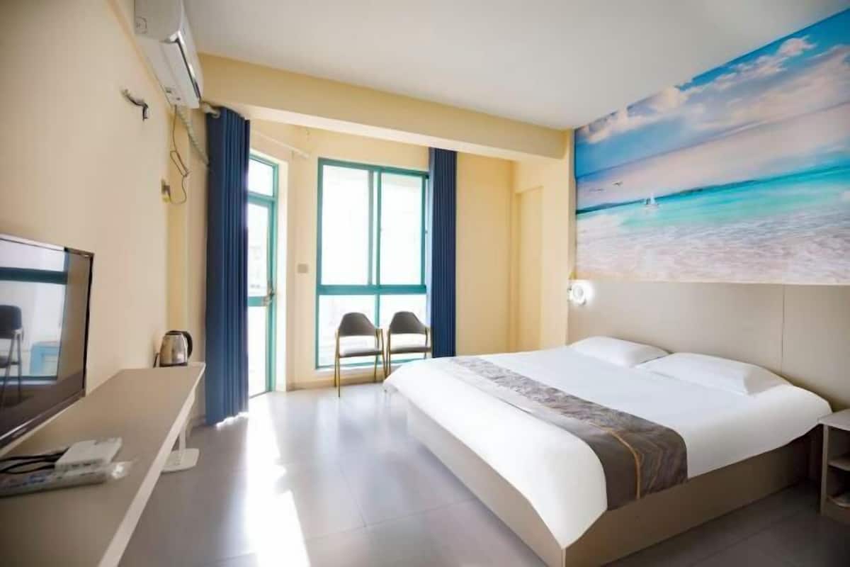 A hotel room with a bed, a desk with a computer, a TV, and a painting of a beach scene.